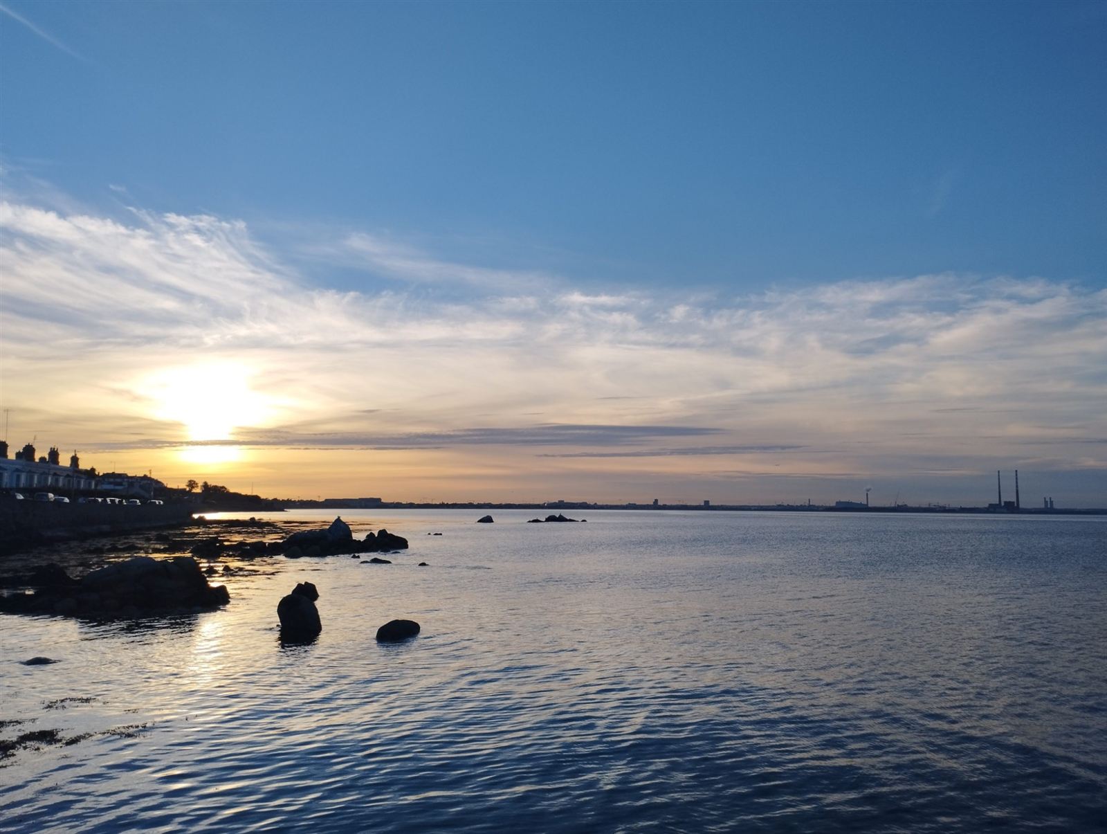 Calm water on Dublin Bay at sunset, with dark rocks in the foreground and the Poolbeg chimneys on the far horizon.