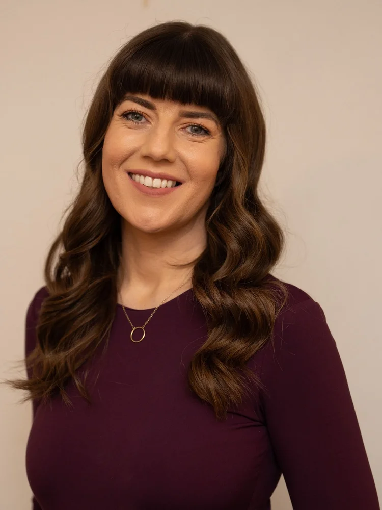 Muireann Banks, souriante, aux longs cheveux bruns ondulés, portant un haut prune foncé.