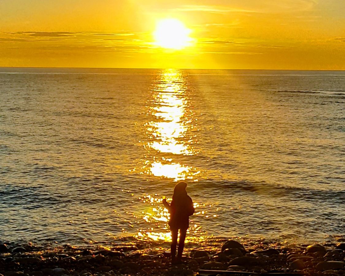 A person stands alone at the water’s edge on a stony beach, looking out at the sun low over the sea.